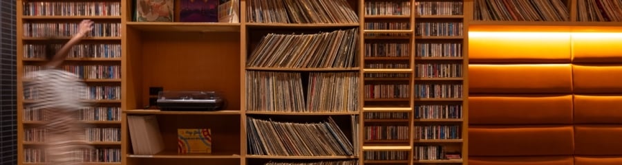 A bookshelf filled with vinyl records and cassettes. A record player is on display, and a velvet chair is in the foreground.